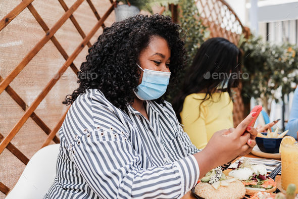 Happy multiracial friends using mobile phones at brunch restaurant while wearing masks