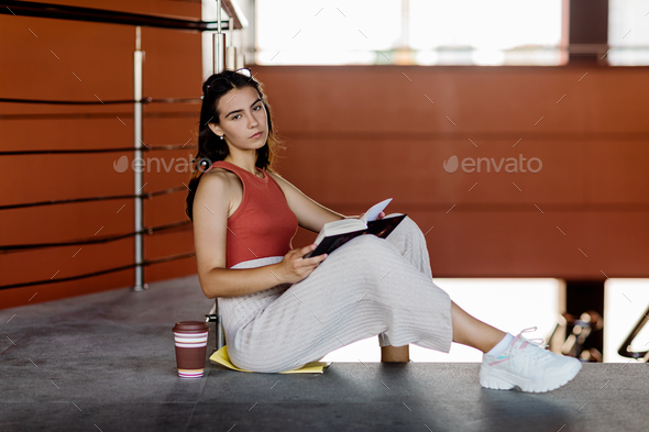 A pretty student girl with a textbook and a cup of coffee is sitting on the stairs - Stock Photo - Images