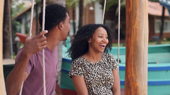 Happy Young Couple Sitting on a Swing at the Beach Laughing and Kissing alt