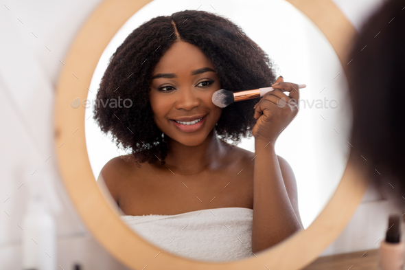 Pretty black lady applying makeup near mirror, using decorative cosmetics indoors