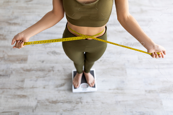 Closeup of Indian female measuring waist, standing on scales, checking weight loss result, measuring