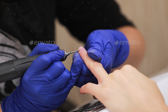 Manicurist in protective gloves is applying electric nail file drill to manicure on female fingers.