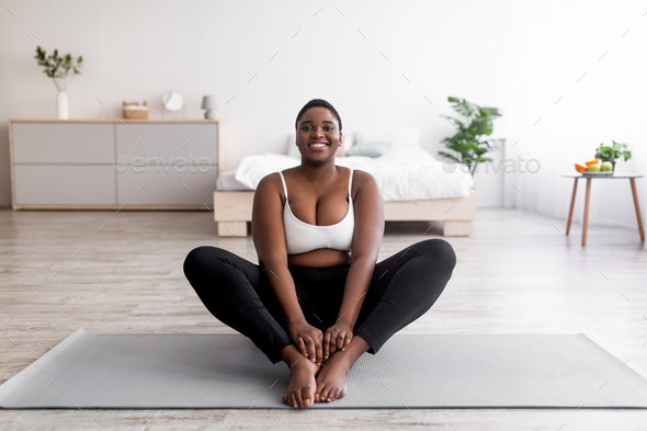 Cheerful plus size black woman doing yoga on sports mat at home