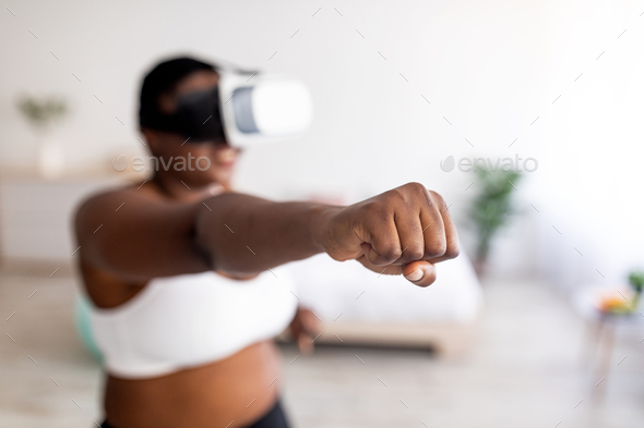 Virtual training. Overweight black lady in VR glasses using fighting simulator, making punch at home
