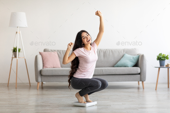 Overjoyed Indian woman sitting on scales, gesturing YES, excited over result of her weight loss diet