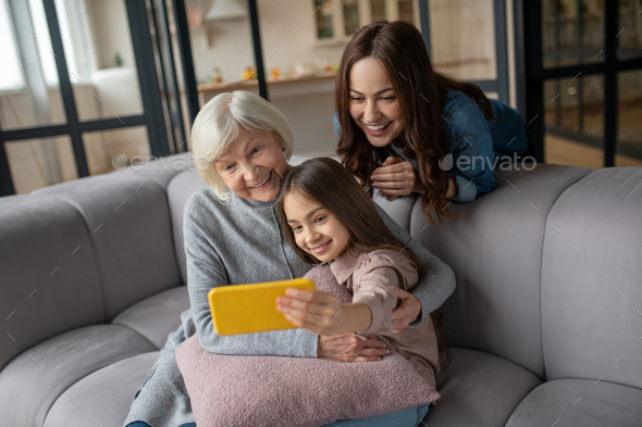 Happy girl, mom, grandmother taking pictures together at home on couch.
