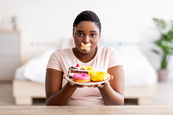 Hungry plus size black woman having weight loss diet, wearing adhesive bandages on mouth, holding