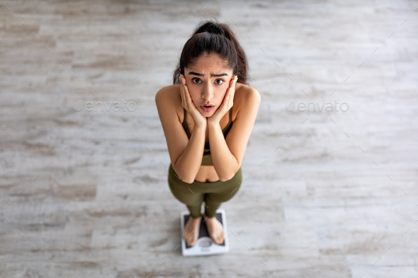 Above view of upset young Indian woman standing on scales, feeling disappointed about her weight