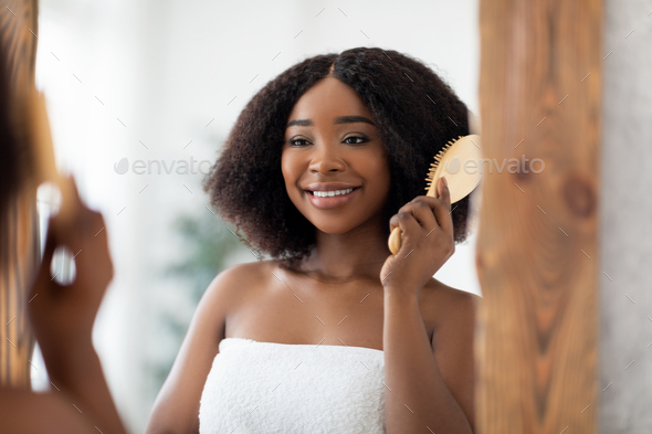 Cheerful Afro woman brushing her curly hair with wooden brush in front of mirror at home