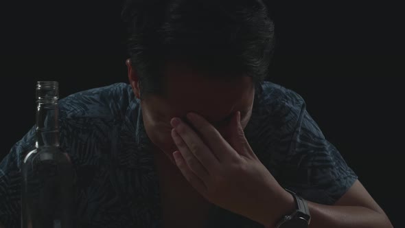 Drunk Asian Man Crying And Pouring Vodka In A Shot Glass Before Drinking In Black Background alt
