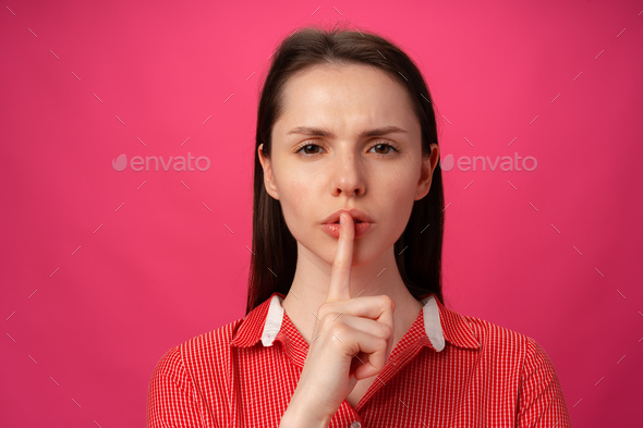 Portrait of woman holding finger on lips to be silent against pink background