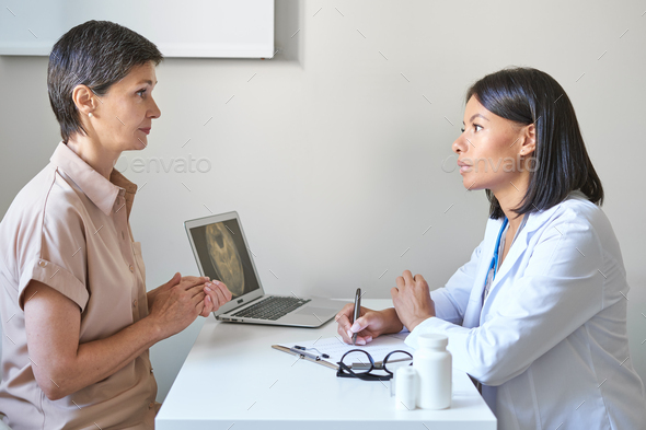 Worried middle-aged woman sitting in clinic cabinet, describing disease symptoms to afro american GP