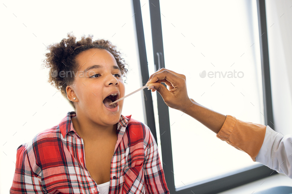Cute likable mixed race teen girl having throat examination with tongue depressor at modern light