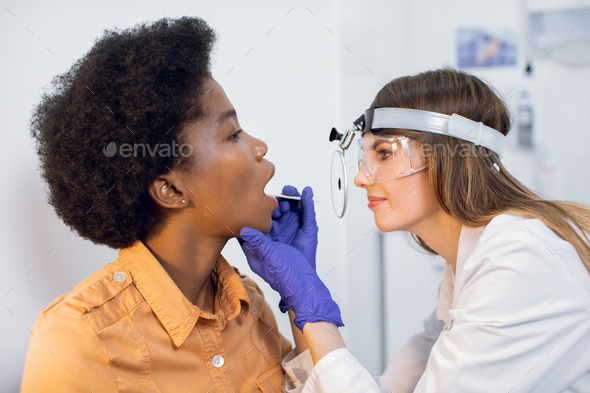 Close up of young afro lady on a doctor\'s examination, while female ENT looks at her throat with