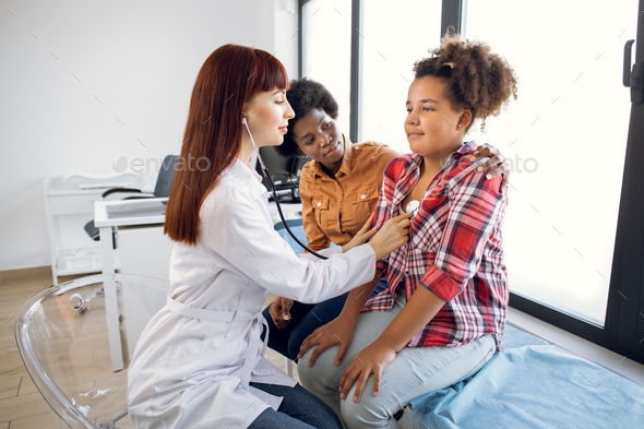 Flu, cough, asthma concept. Female pediatrician doctor listens to lungs of African-American 12-aged