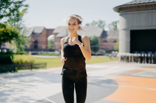 Pleased energetic woman runs quickly on stadium, has healthy fit body listens music