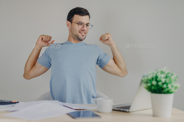 Overworked male stretches at work place, just finishes with work, looks happily into screen
