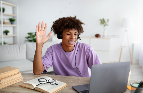 Black teen guy in headphones waving at laptop webcam, speaking to teacher or fellow students, having