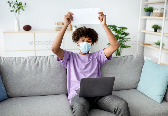 African American youth in face mask showing empty sign with mockup, sitting on couch with laptop