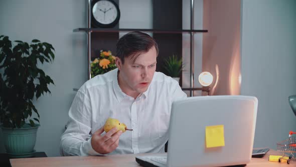 A man eats a pear in his workplace and works in front of a computer
