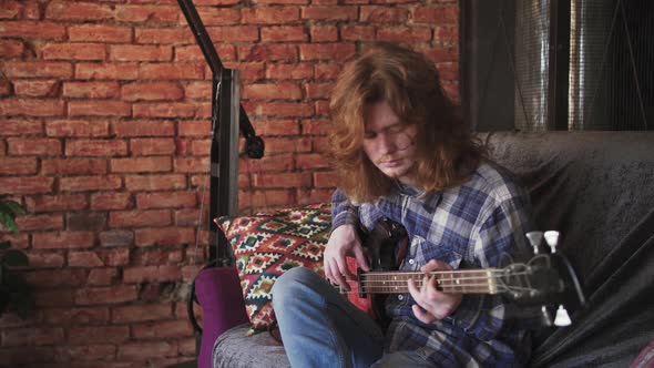 Portrait of Young Man Musician with Long Red Hair Playing Bass Guitar at Home Sitting on Sofa alt