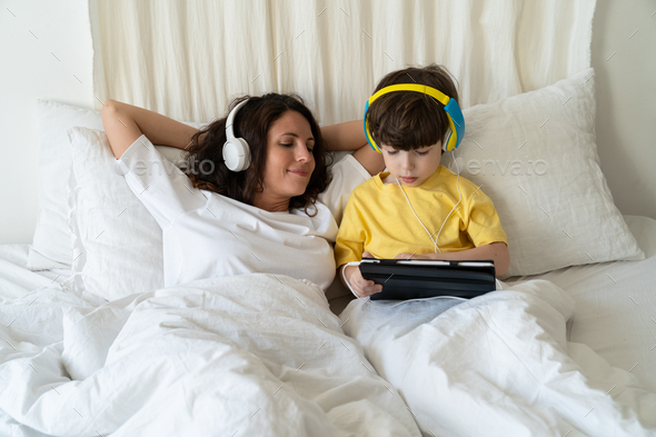Family morning in bedroom: mom in headphones look at little kid son play games on tablet computer