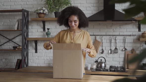 Happy Cheerful Frican American Girl Gets the Delivery Box. Happy Box Openning in a Kitchen alt