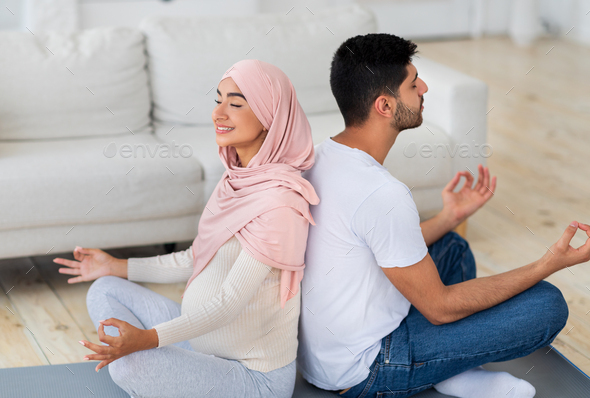 Yoga for pregnancy. Happy muslim spouses meditating together at home, sitting back to back and