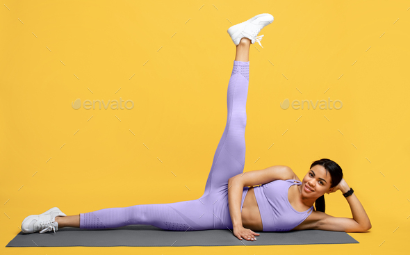 Young sporty black lady warming up and lifting leg up, exercising on mat over yellow background
