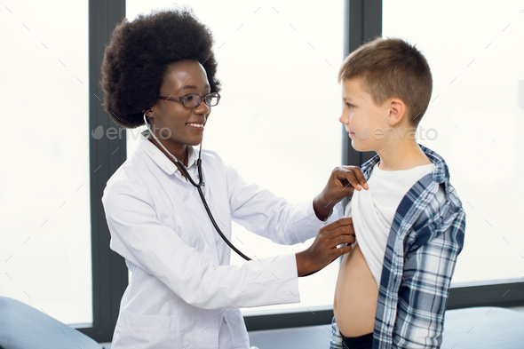 Cute likable Caucasian teen boy and joyful African woman doctor, during doctors checkup. Pediatrist