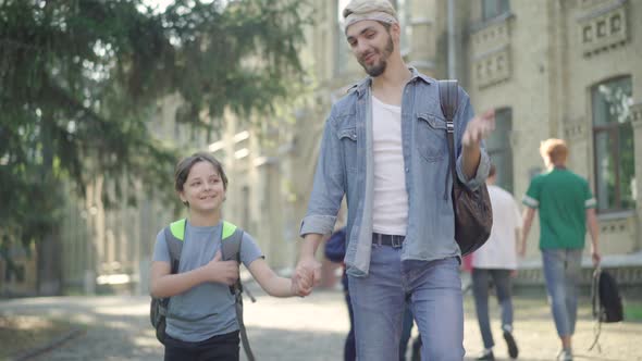 Portrait of Positive Caucasian Brothers Walking Along Schoolyard in Sunlight. Smiling Young Man and alt