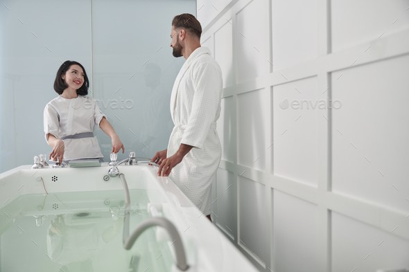 Woman in uniform shows hydro massage bath to man guest in modern salon