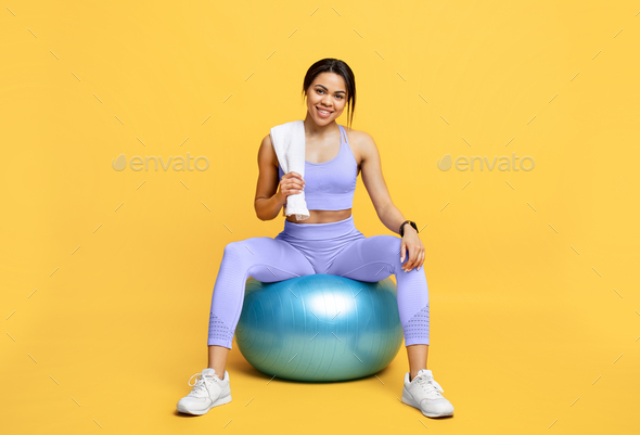 Equipment for training. Slim black lady in sports uniform holding towel and sitting on fitball over