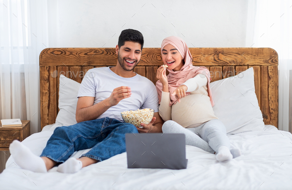 Funny comedy. Happy muslim man and pregnant wife watching film on laptop  computer, eating popcorn Stock Photo by Prostock-studio