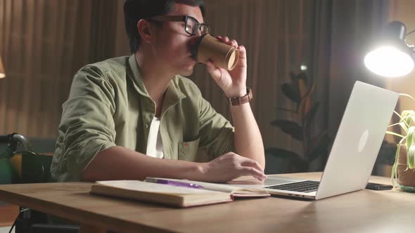 Asian Man Sitting In A Wheelchair While Working On Laptop Computer And Drinking Coffee At Home alt