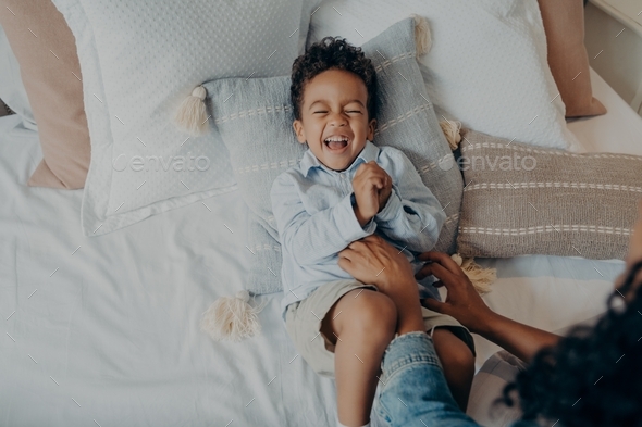 Adorable little son lying on pillows in bed and laughing while mom tickling him