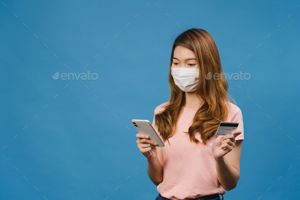 Young Asia lady wearing medical face mask using phone and credit bank card with positive expression.