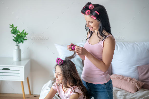 Dark-haired mom with hair curlers curling hair to her daughter