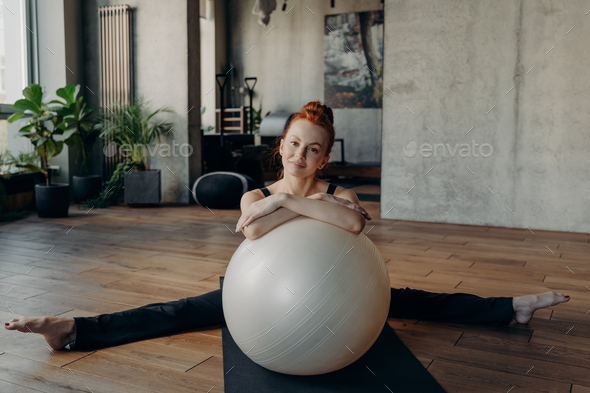 Young athletic redhead woman sits in twine with fitness ball