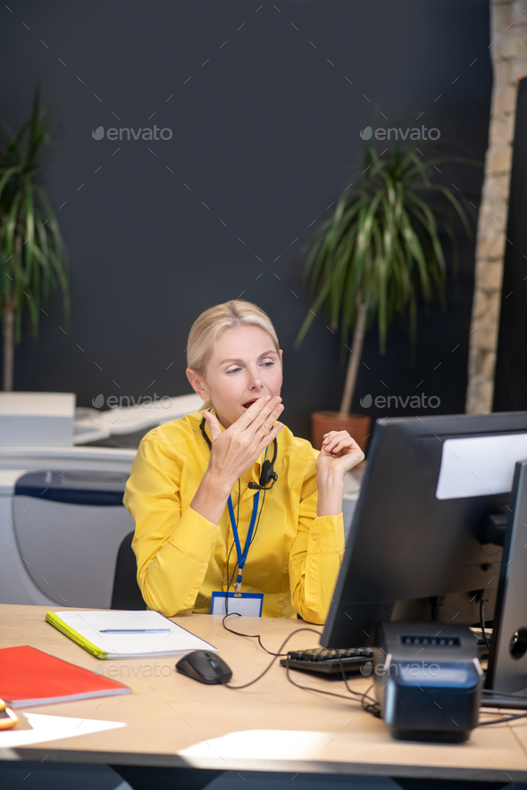 Sleepy blonde woman sitting in headphones at desk, yawning