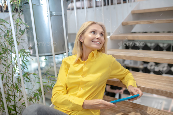 Blonde woman sitting on stairs, holding blue tablet, smiling, looking sideways