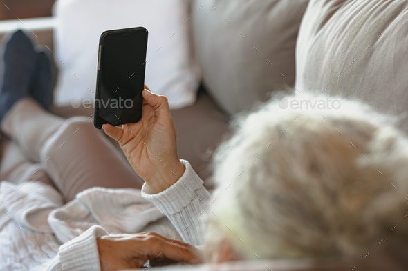 Senior caucasian woman laying and using smartphone in the modern living room - Stock Photo - Images
