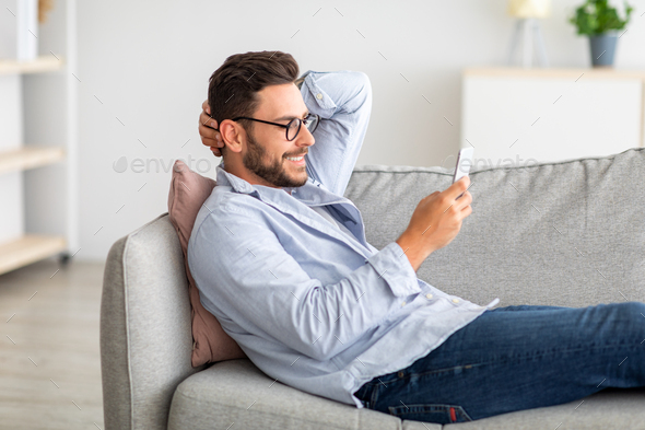 Cool gadget and app. Young smiling guy holding mobile phone, typing sms message, resting on sofa