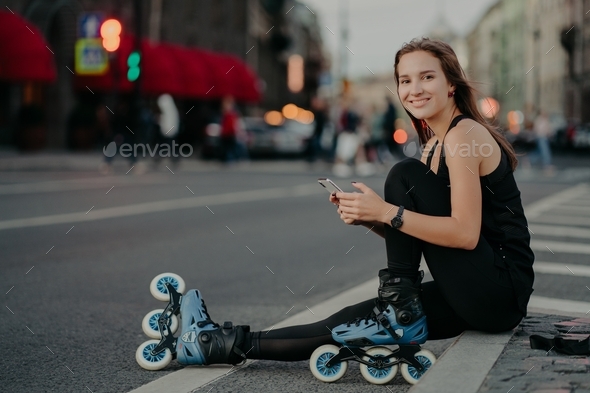 Active slim woman being in good physical shape rides rollerblades uses smartphone