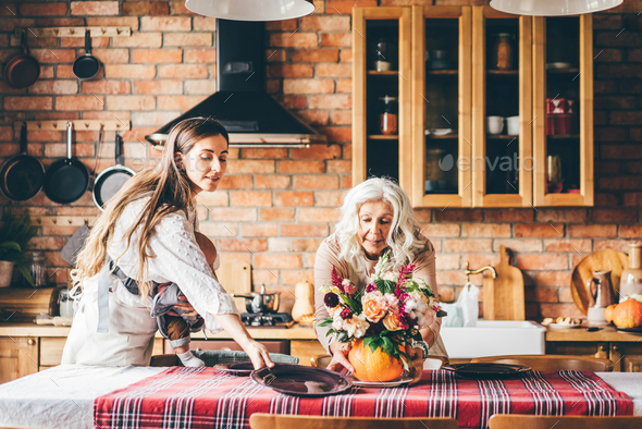 woman holds baby in sling bag and helps aged mother serving large table with autumn flower bouquet