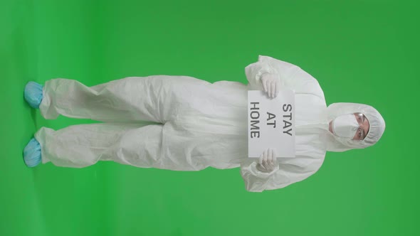 Man Wearing Protective Uniform PPE And Holding Stay At Home Sign In The Green Screen Studio alt