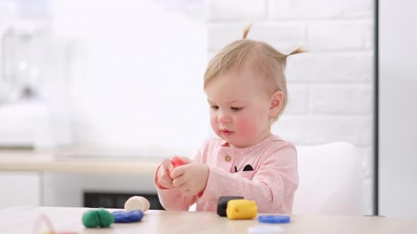 Cute Little Girl with Funny Ponytails on Head Sitting in Kitchen and Playing Multicolored Plasticine alt