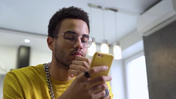 A Young Man Wearing Glasses Reads Messages on a Smartphone at Home alt