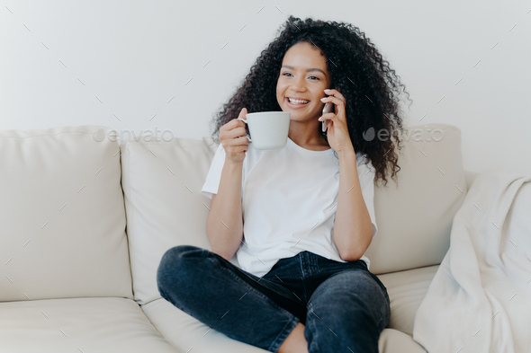 Cheerful Afro American woman has coffee break in living room, sits on couch, calls friend smartphone