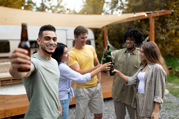 Arab guy and diverse friends making toast with beer bottles, having party with alcohol drinks on